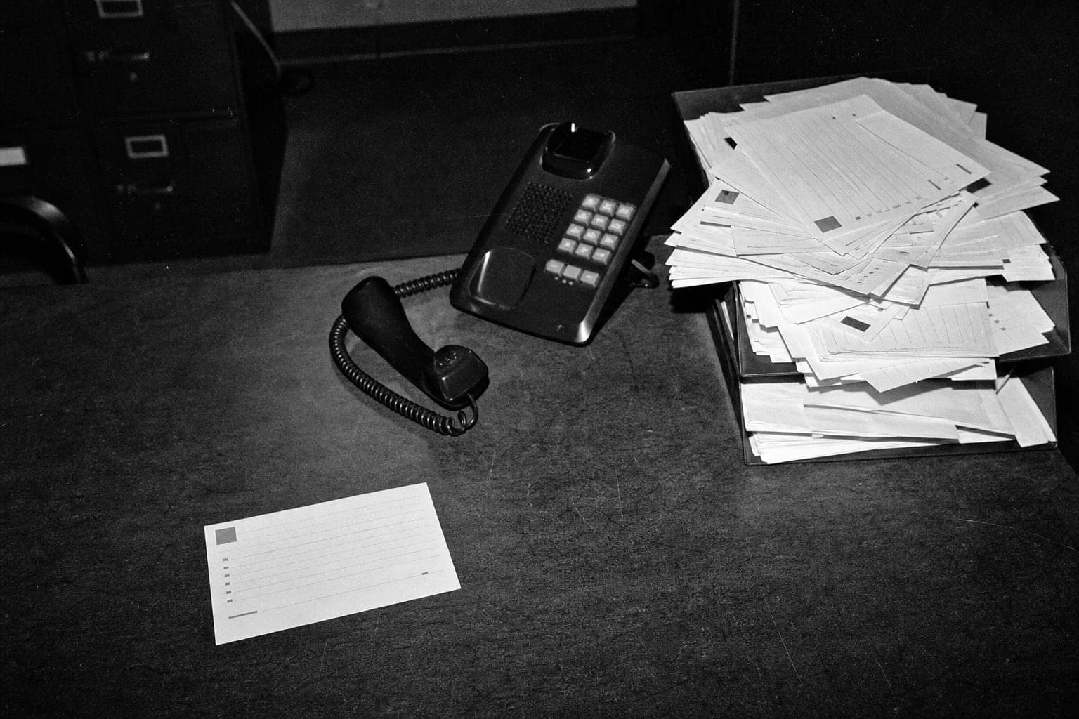 An unattended desk phone beside an overflowing inbox tray of inquiry sheets, with one overlooked sheet isolated in the foreground.